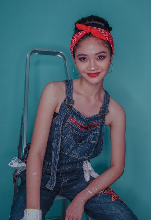 Free Smiling woman in denim overalls sitting on a ladder against a teal background. Stock Photo
