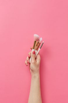 A hand holding makeup brushes against a vibrant pink background, showcasing beauty essentials.