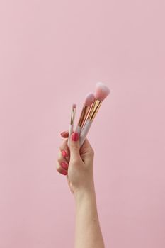 Aesthetic shot of a hand holding elegant makeup brushes against a light pink backdrop.