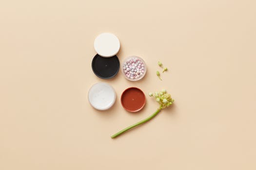 Artistic flat lay of makeup containers, powder balls, and flower buds on a soft yellow surface.