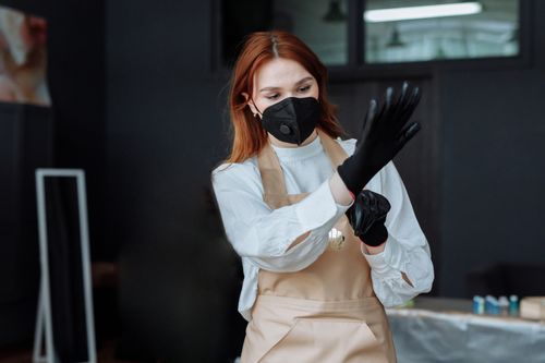 Free A woman wearing a mask and gloves adjusts her attire, ready for work in a professional indoor setting. Stock Photo