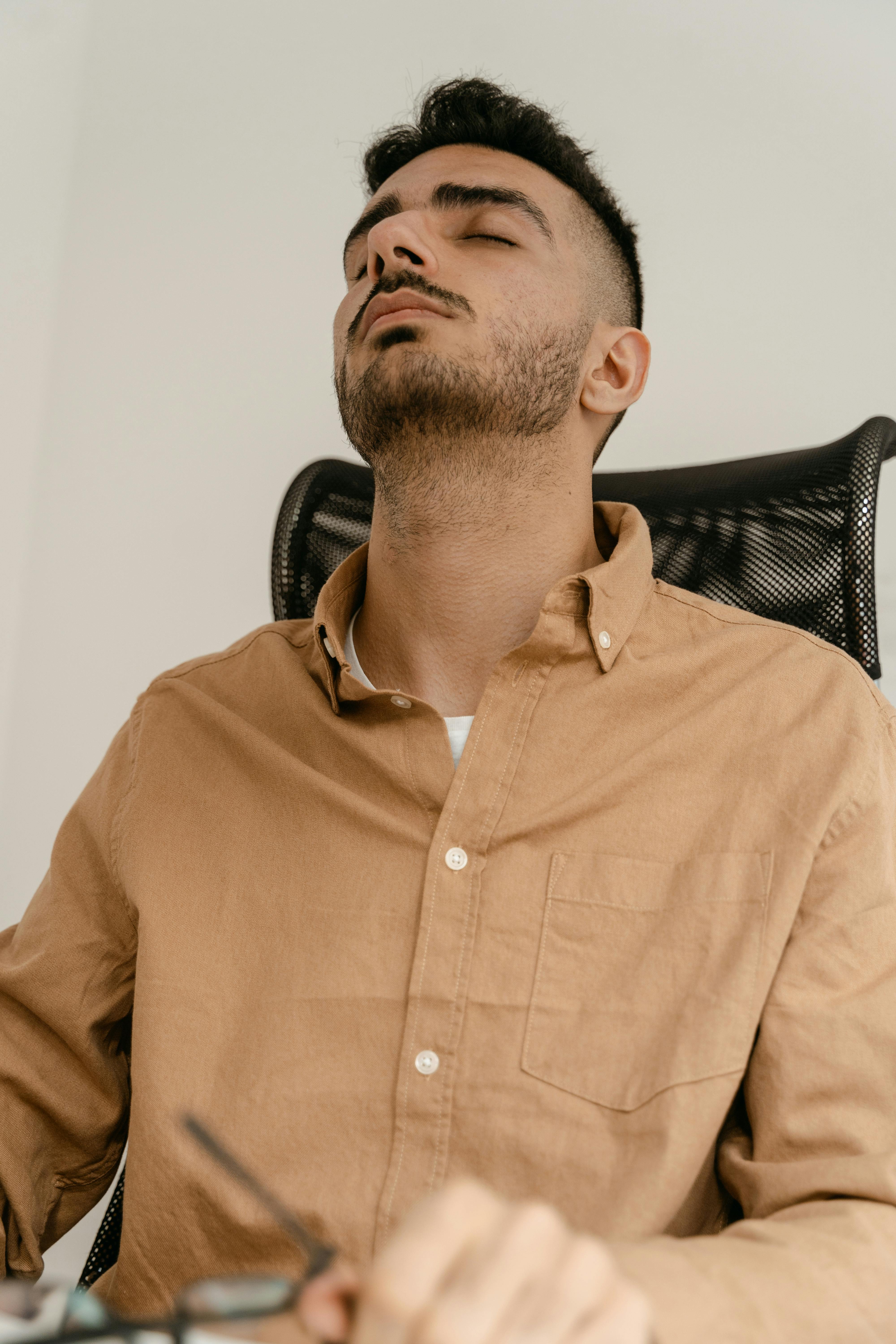 Tired man in brown shirt with closed eyes, appearing stressed and weary in office setting.