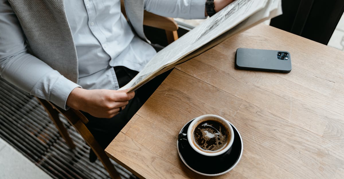 An adult male enjoying his coffee while reading a newspaper at a cafe with his smartphone nearby.