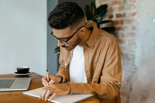 Free A man wearing glasses and a brown shirt writes notes at a modern workspace with a laptop. Stock Photo
