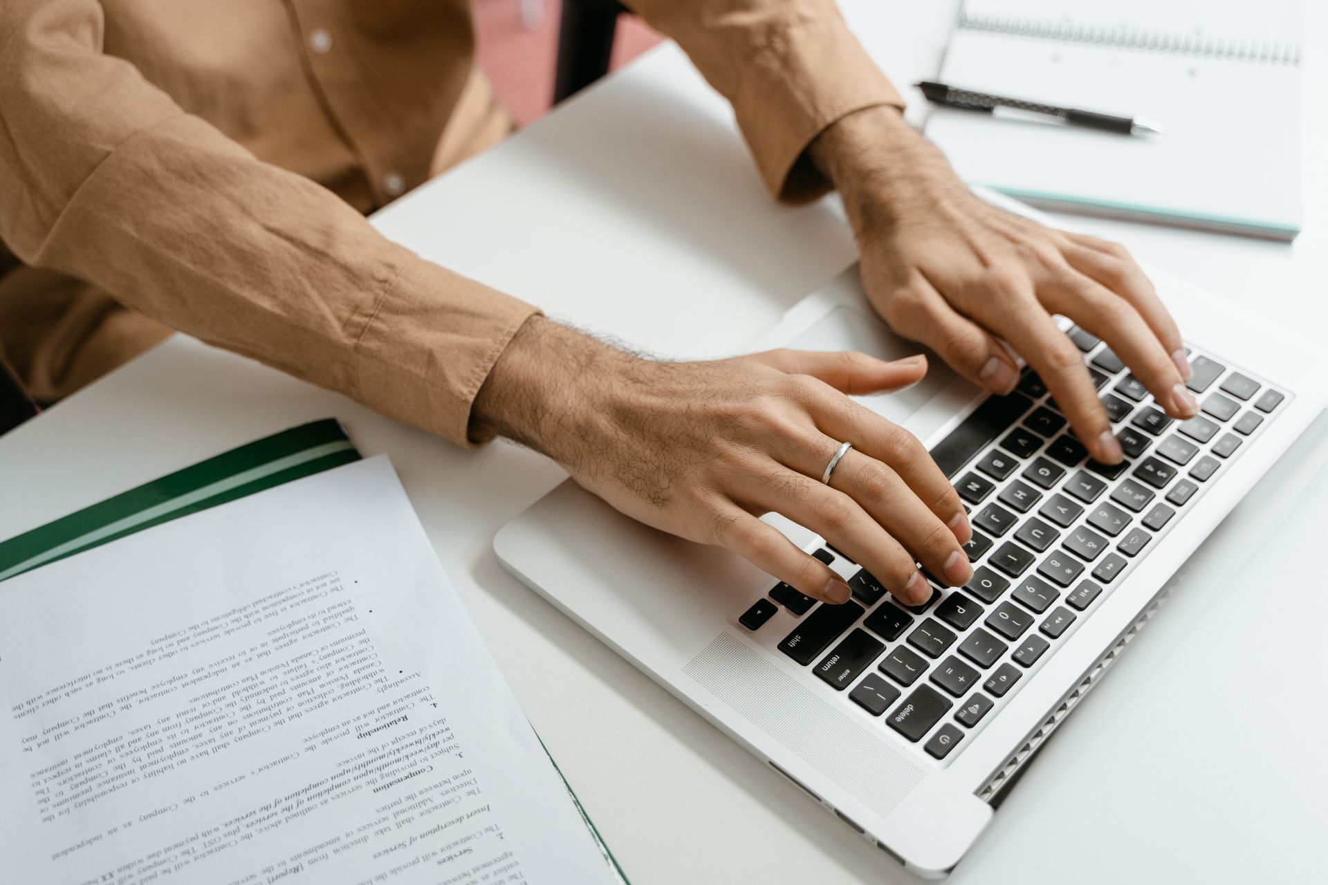 Close-up of hands typing on a laptop keyboard with documents on a desk, brown long sleeves visible.