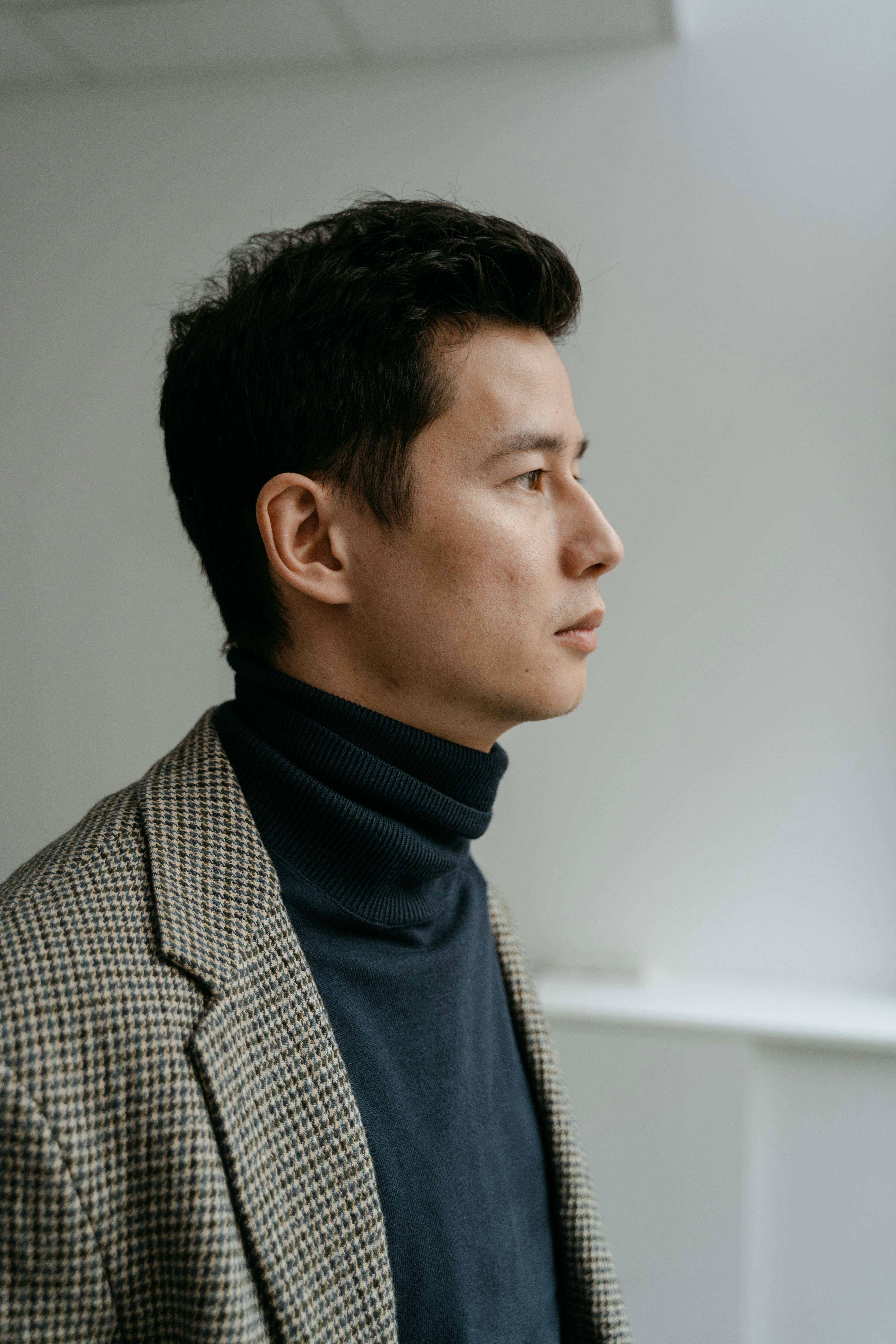 Side profile of a man wearing a checkered coat and navy turtleneck against a simple background.
