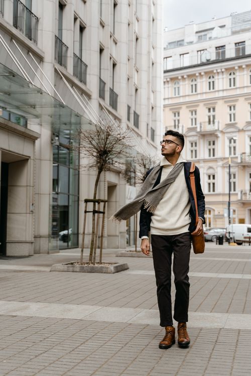 Free Adult man walking on city sidewalk, wearing eyeglasses and scarf, showcasing urban fashion. Stock Photo