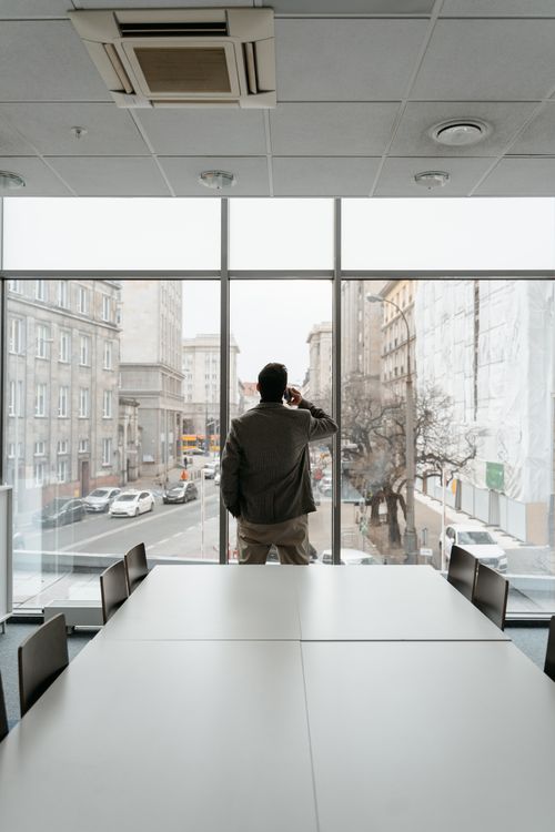 Free Businessman standing in a modern office, talking on the phone while looking out the window onto a city street. Stock Photo