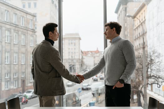 Two businessmen shaking hands in an urban office with city view through large windows.