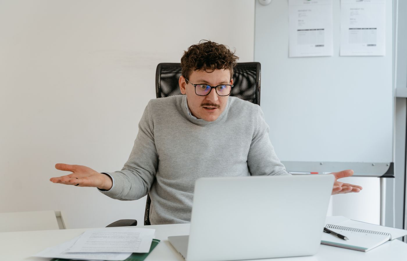 A man in a gray sweater and glasses gesturing at his laptop screen during a video call in an office setting.