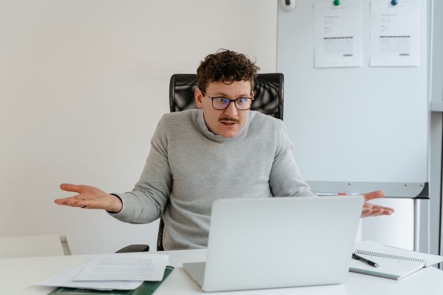 A man in a gray sweater and glasses gesturing at his laptop screen during a video call in an office setting.