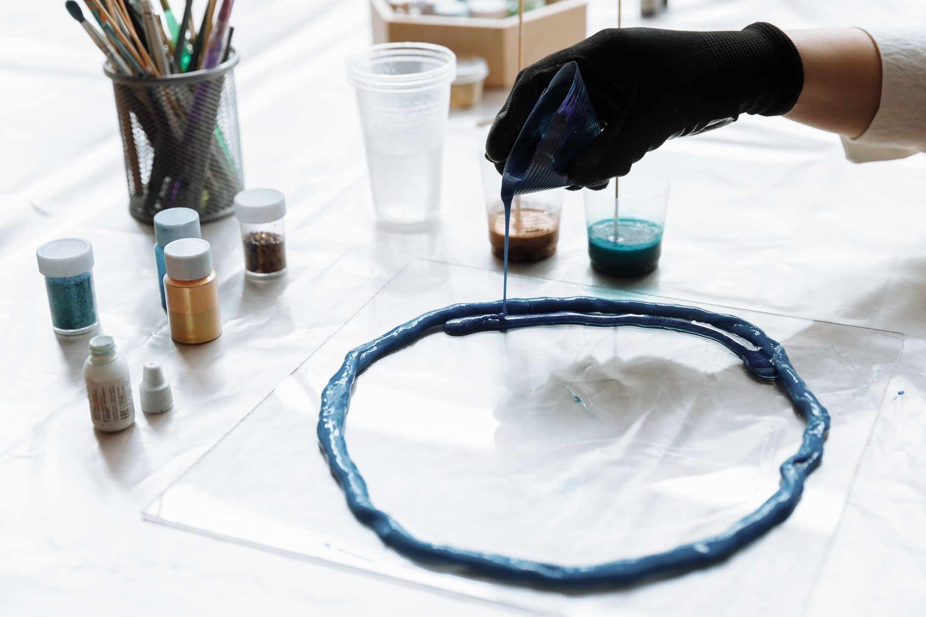 Close-up of gloved hand pouring blue resin onto a surface with art supplies.