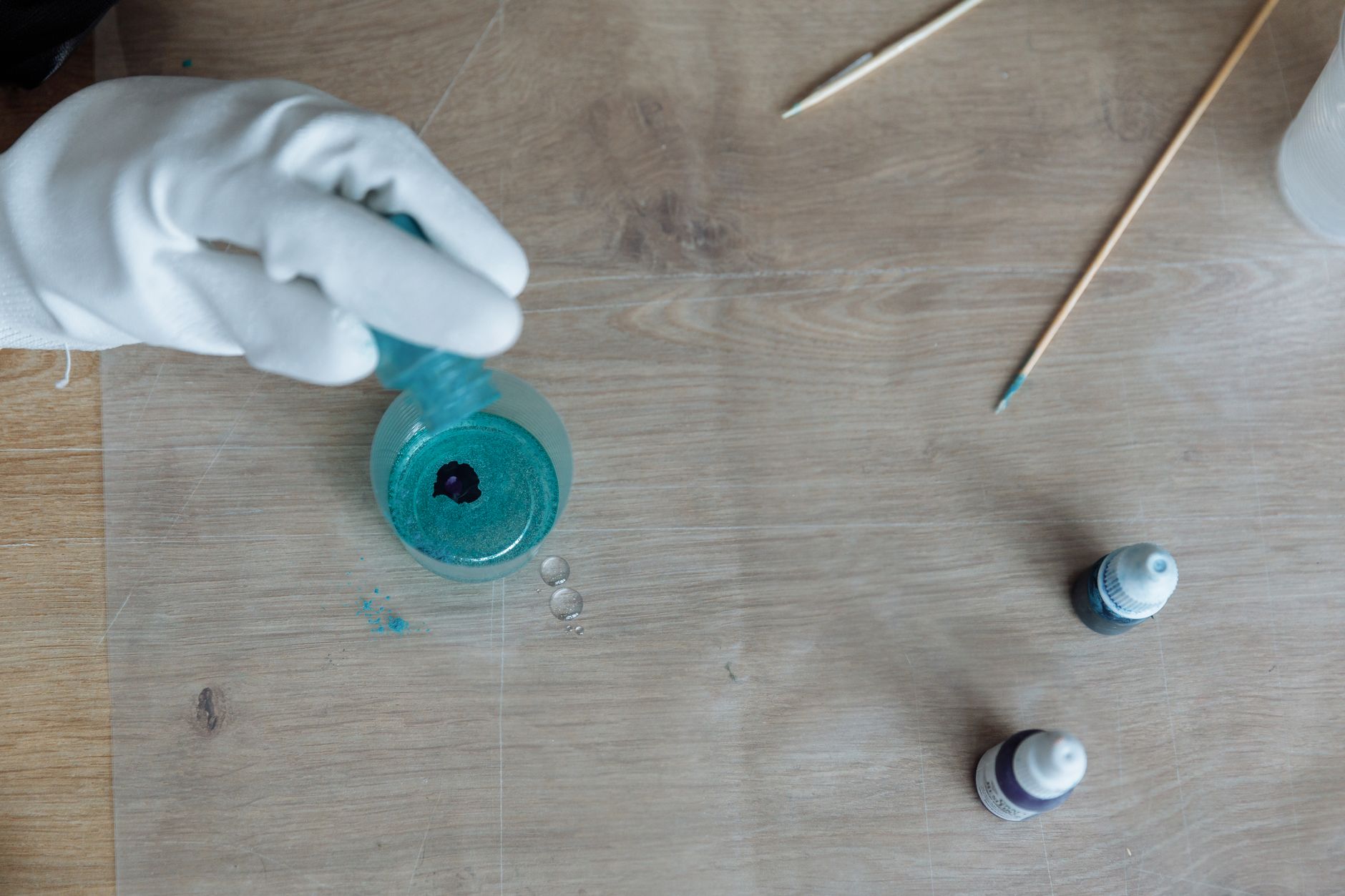 A gloved hand pouring blue resin into a container on a wooden table, creating art.