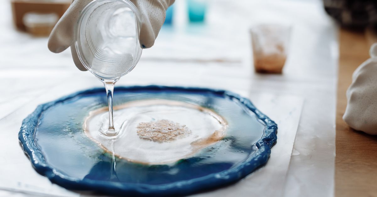 A gloved hand pouring clear resin into a blue geode-shaped mould on a bright work table