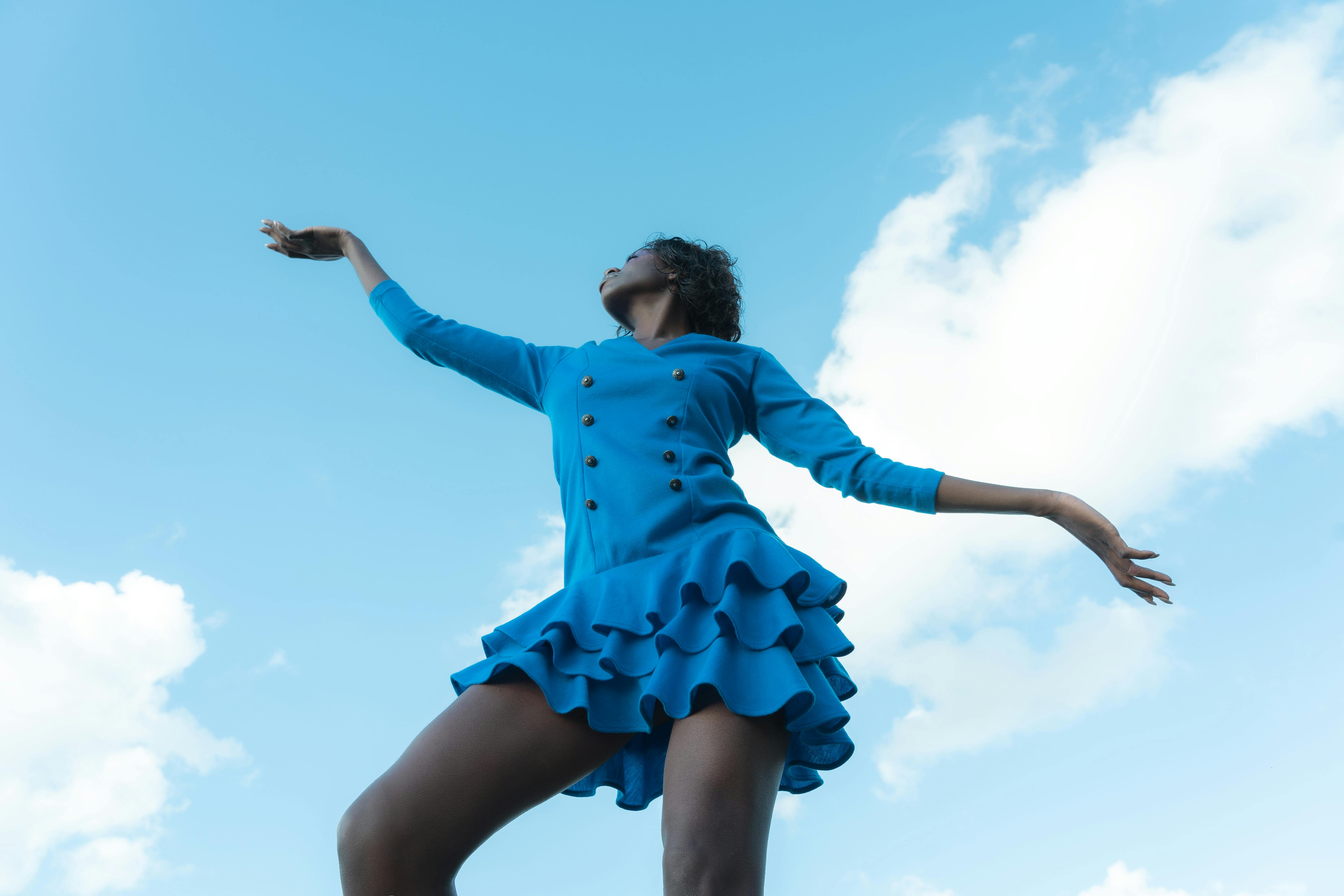 Woman in a blue dress striking a graceful pose with a sky background, showcasing elegance.