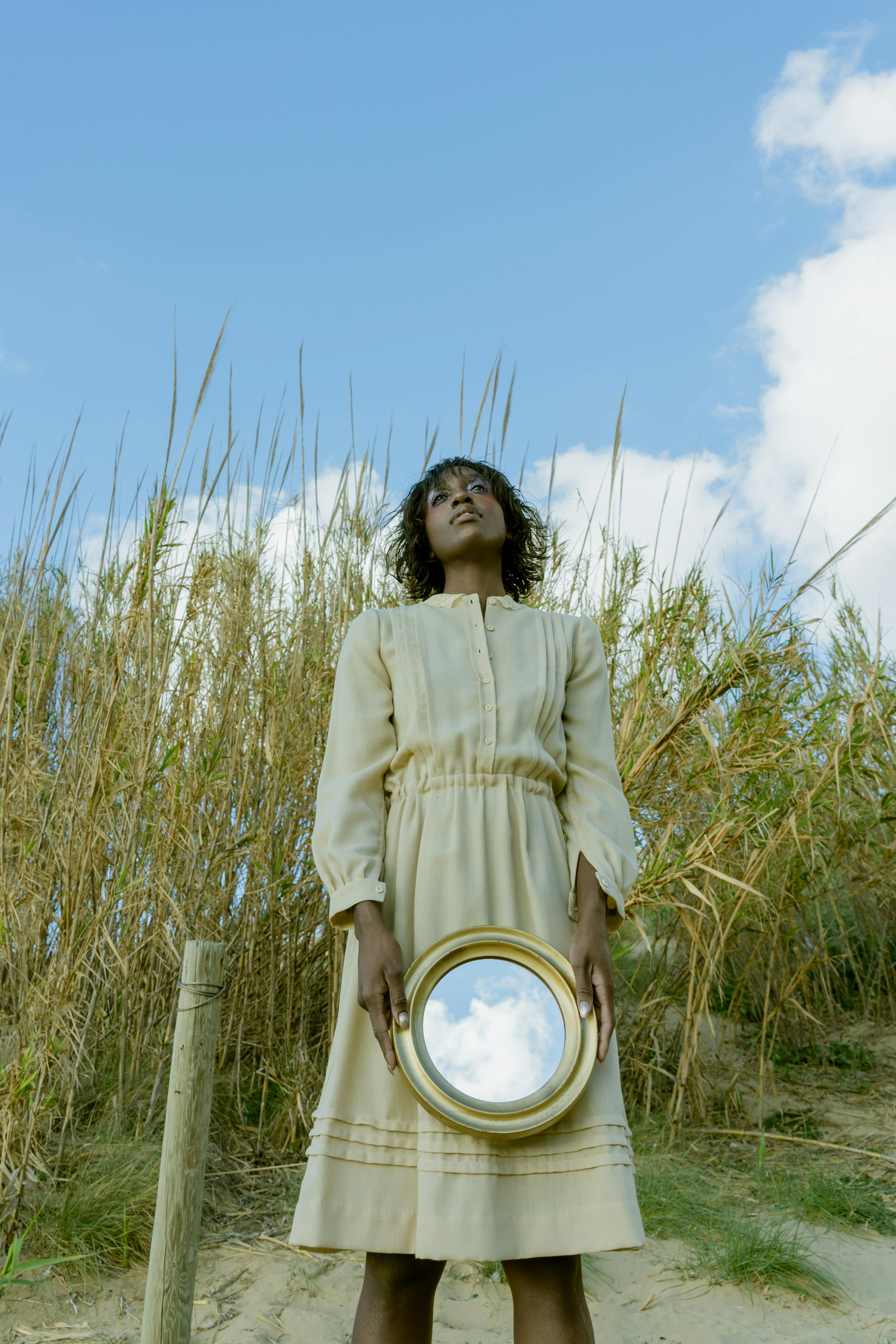 Artistic portrait of a woman holding a mirror in a natural setting with grass and sky.
