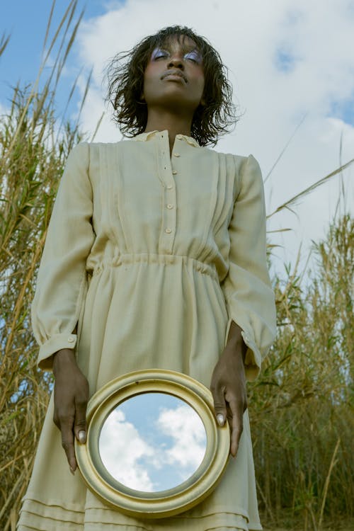 Woman in a Dress Standing in a Field with her Eyes Closed Holding a Mirror