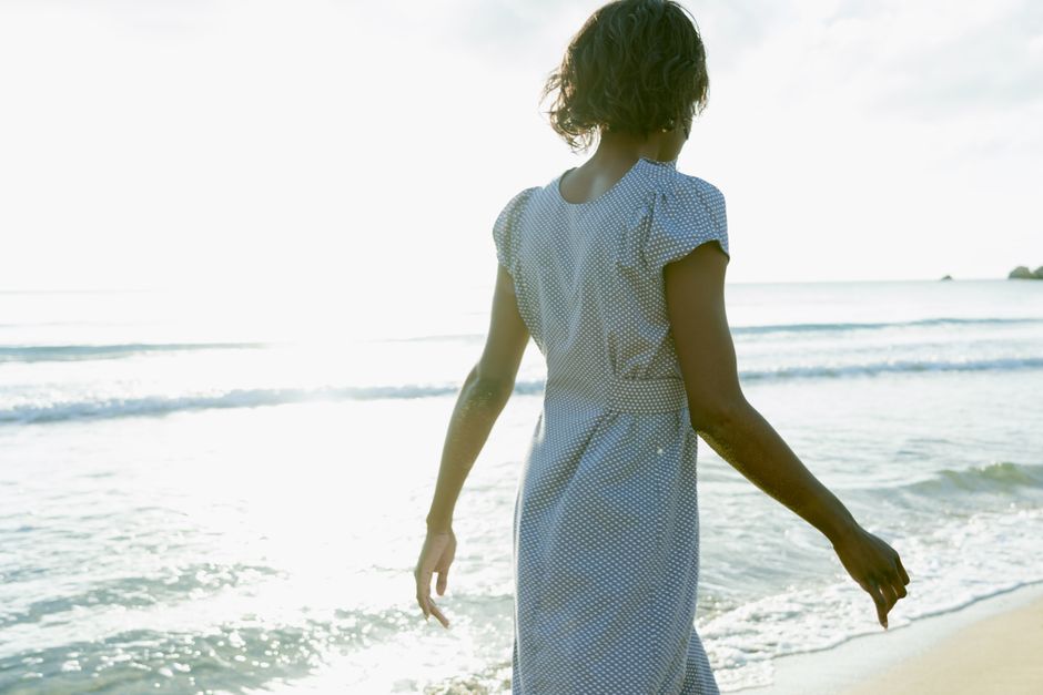 A woman in a polka dot dress walks along a sunlit beach shoreline.