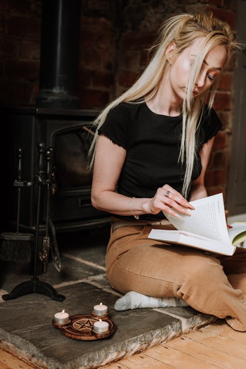 Free Serious blond female looking through pages of book during spiritual practice with candles and pentagram near fireplace Stock Photo