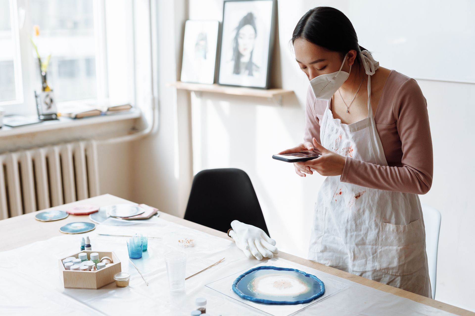 Woman in apron with face mask capturing her resin art creation using a smartphone indoors.