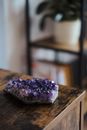 Natural uncut amethyst with violet crystals placed on wooden cupboard in light room with potted plant on blurred background at home