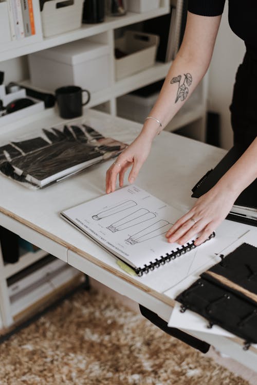 Unrecognizable tattooed female dressmaker standing at table with sketchbook with sketches of collection of clothes while working in modern studio