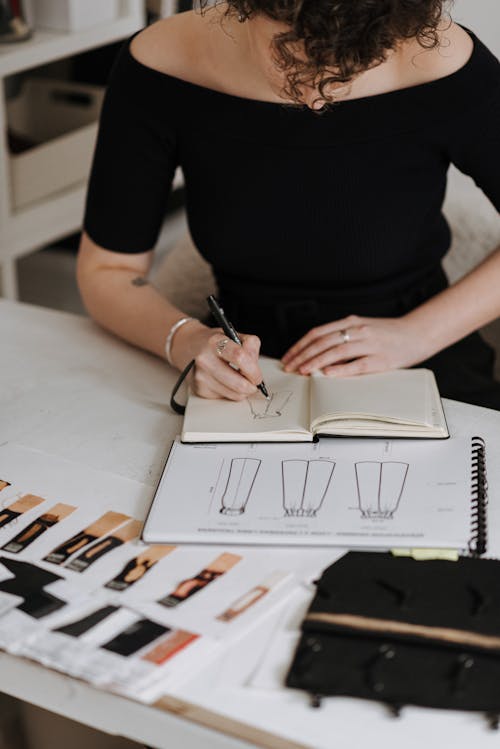 Anonymous female designer drawing sketch of new clothes while sitting at table with samples of fabrics during work in studio