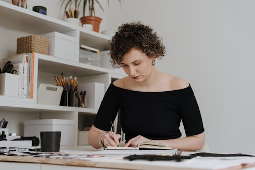 Concentrated talented dressmaker in black outfit drawing draft of clothes while sitting at table in modern workshop with various supplies