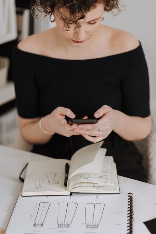 Free Crop female dressmaker text messaging on cellphone while sitting at table with sketches of clothes during work in modern studio Stock Photo