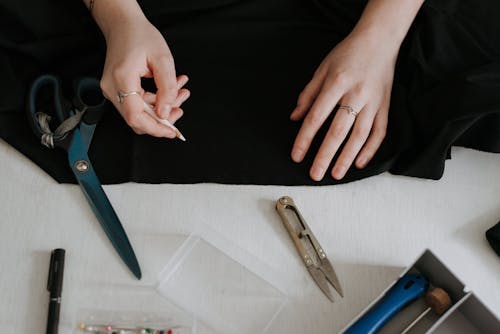 From above of professional female seamstress with pencil preparing to draw outline on black textile while standing at table with scissors and nippers
