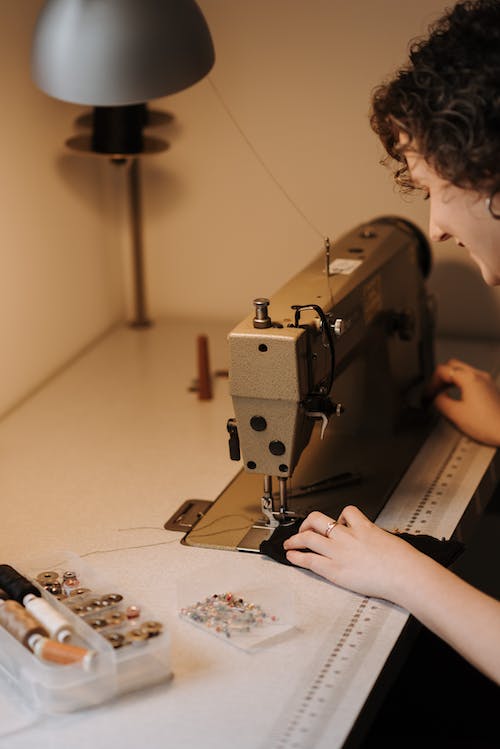 Side view of content skilled female dressmaker using sewing machine at table with sewing supplies while working in professional workshop