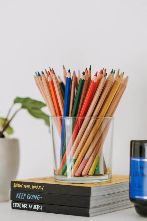 Free Set of various multicolored pencils in glass cup placed on stack of books in light room with green plant on white background Stock Photo