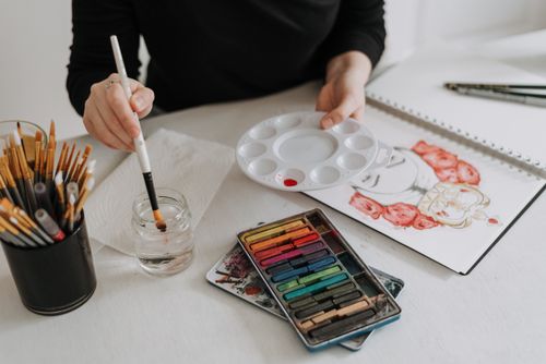 Free Unrecognizable painter with palette and paintbrush sitting at table with sketchbook and art supplies while painting in modern light studio Stock Photo