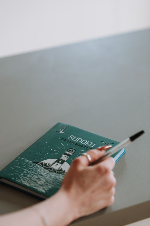 Free Close-up of a hand holding a pen beside a blue Sudoku book on a grey table, ready for a puzzle challenge. Stock Photo