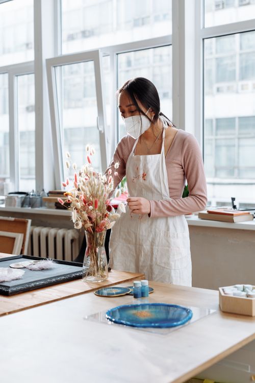 Free Woman with face mask arranging dried flowers in a vase, engaged in arts and crafts at an indoor studio. Stock Photo