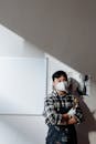 A Man in Plaid Long Sleeves Leaning on the Wall with Whiteboard while Wearing Face Mask