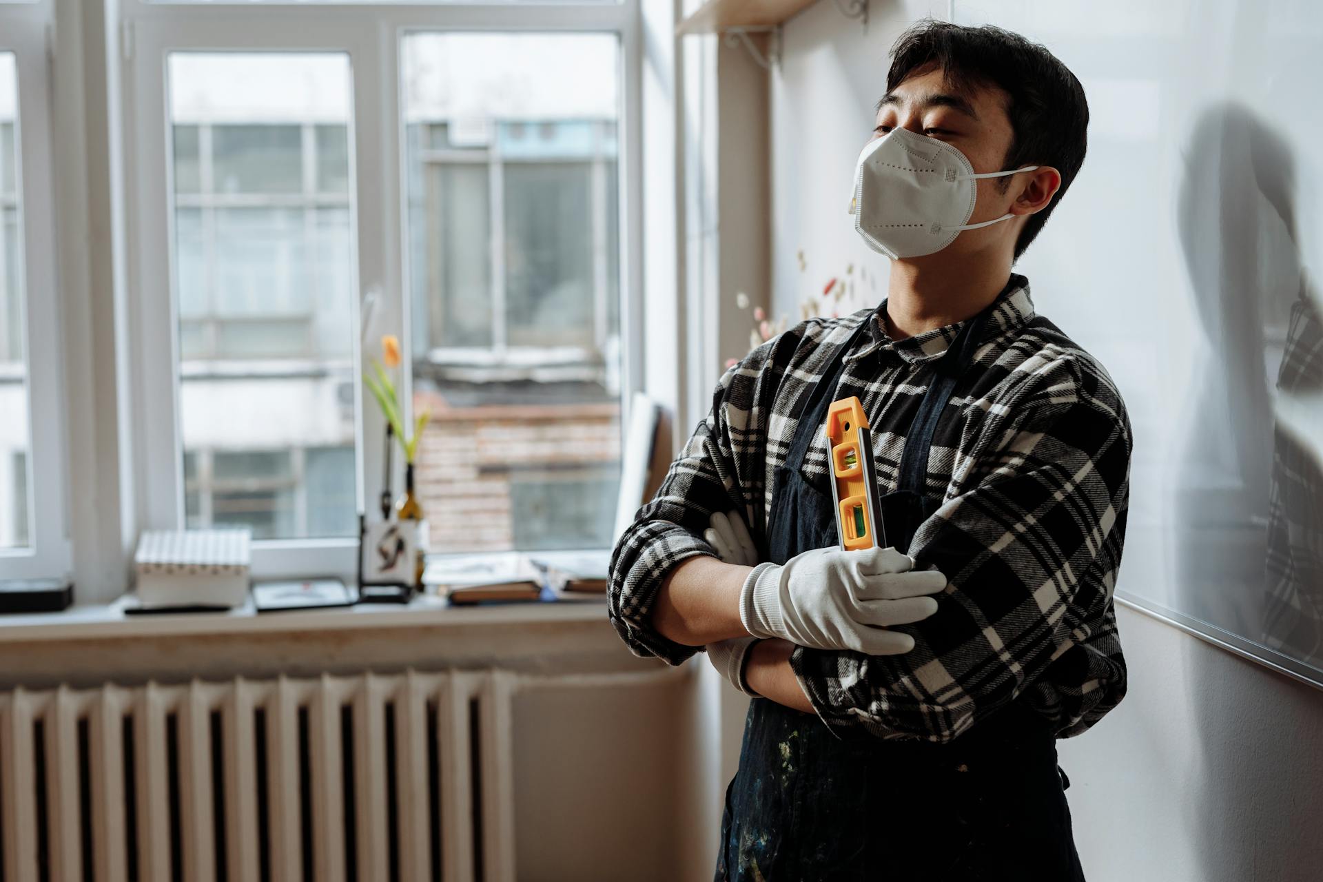 A man in a plaid shirt and face mask stands indoors holding a level tool.