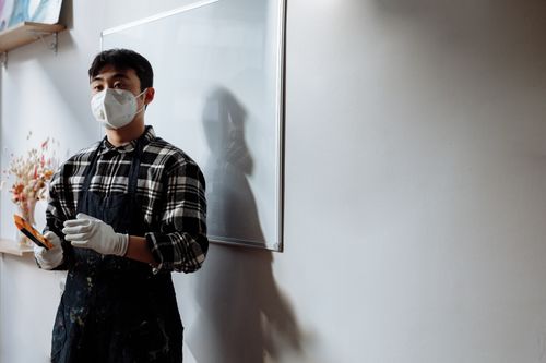 Free An artist wearing a face mask and apron holds brushes in a well-lit room with a whiteboard. Stock Photo