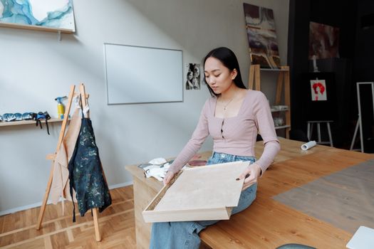 Asian woman unpacks a wooden box in a contemporary art studio, surrounded by creative materials.