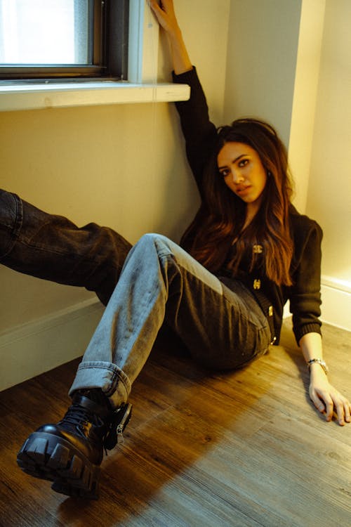 Rebellious young ethnic female millennial with long dark hair and makeup in fashionable outfit and boots sitting on floor with raised leg near window and looking at camera