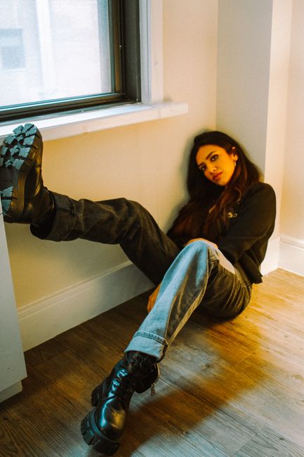 Full body of eccentric young ethnic lady in trendy outfit and boots sitting on parquet floor near window and looking at camera