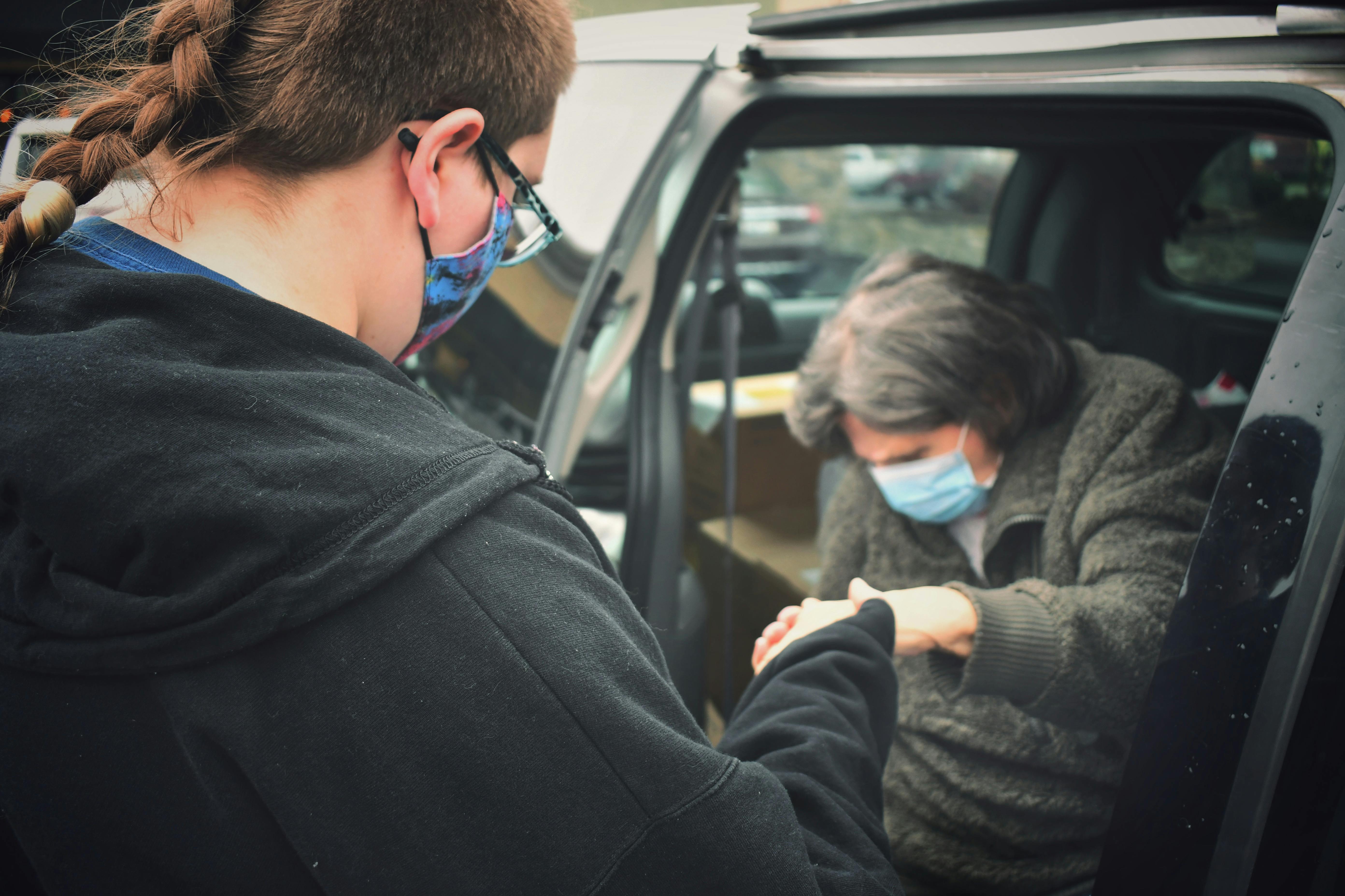 A person assists an elderly man wearing a mask by their car, symbolizing care during the pandemic.