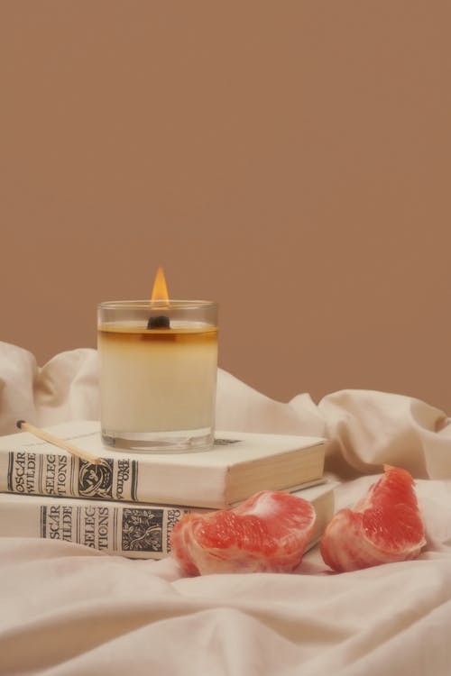 Flaming wax candle placed on books near pieces of grapefruit on creased fabric against beige background