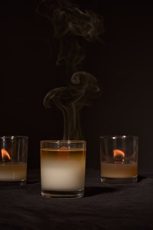 Burning aroma candles in glass holders emitting smoke placed against black background in studio