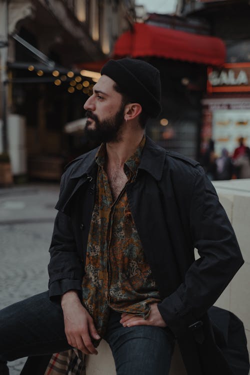 Confident ethnic male with beard in hat looking away while sitting on blocks on paved walkway against old fashioned residential houses in city