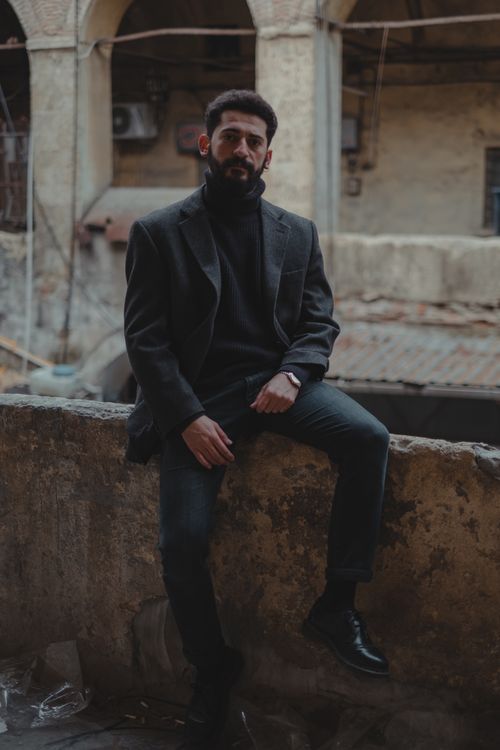 Free Full body of serious bearded ethnic male in elegant wear looking at camera while sitting on concrete barrier of aged demolished building Stock Photo
