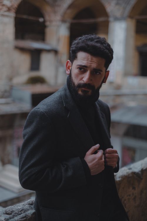 Free Serious ethnic male with beard wearing formal clothes looking away while standing in destroyed building with arched passages on blurred background Stock Photo