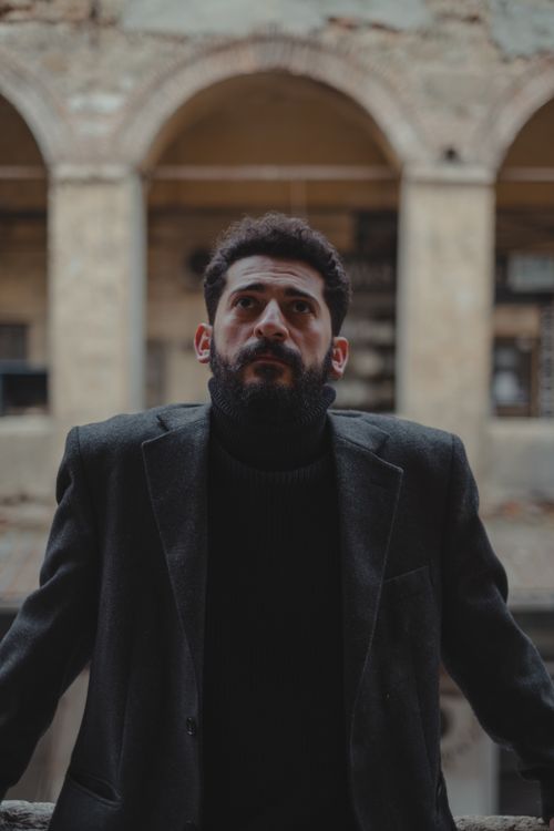 Free A man in a dark suit gazes upward against a rustic urban backdrop with arches. Stock Photo