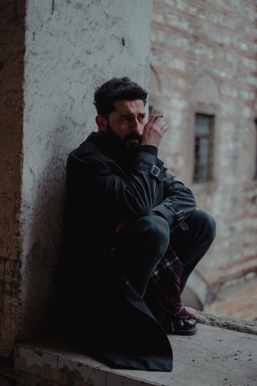 Free Full body of serious bearded man smoking while sitting on shabby stone window opening and looking away on blurred background Stock Photo