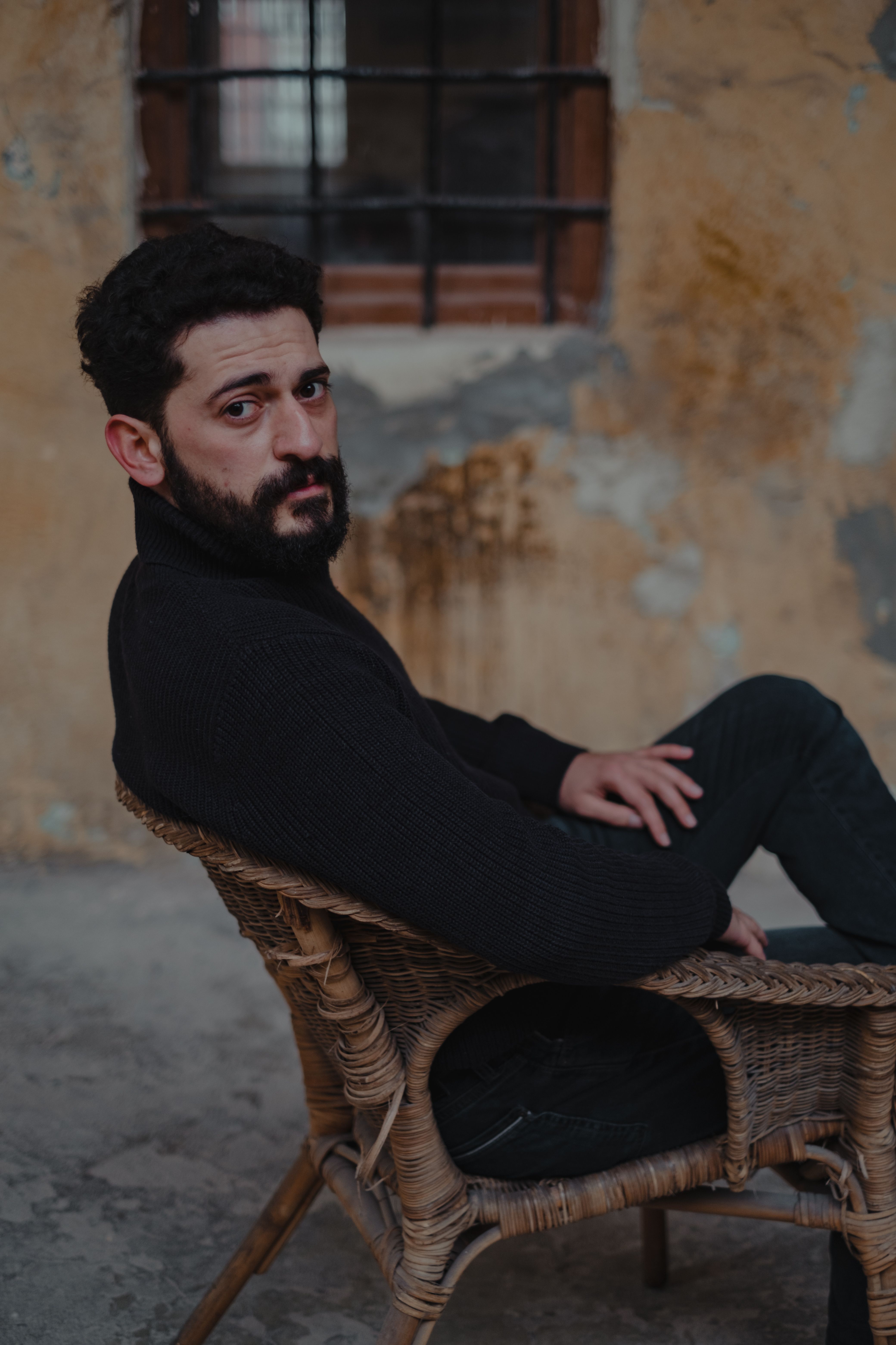 Bearded male in black clothes sitting on wooden chair near shabby wall and looking at camera